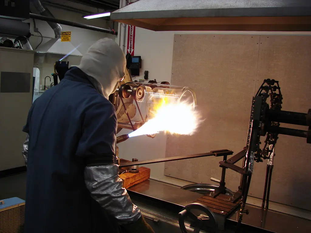 From the very large to the very small, Precision Glassblowing does it all. A glassblower wears a hood and works close to a flame on a large project.
